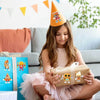 A young girl wearing an orange party hat holds a wrapped DIY pizza sticker craft kit.