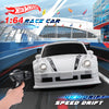 A white 1:64 scale race car is shown on a track with a hand holding a remote control, set against a blurred background.
