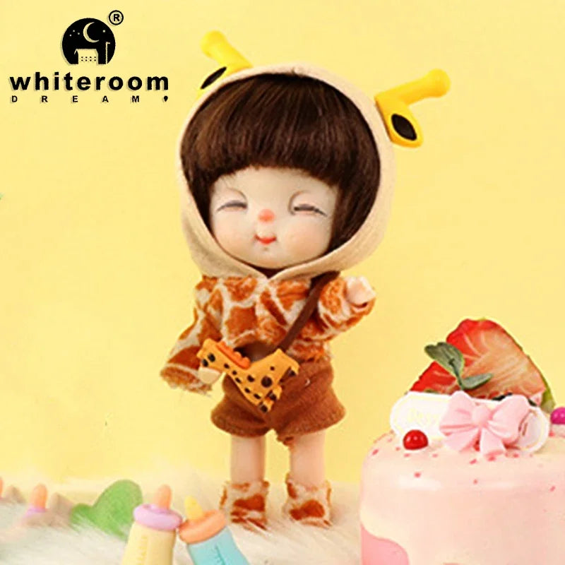A small brown doll with black hair and yellow ears is shown next to a pink cake decorated with fruit and flowers.
