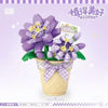 A purple floral bouquet made of building blocks is displayed in a pot with a bow.