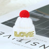 A white knitted hat with a red pom-pom and gold glitter lettering spelling LOVE resting on a dark surface.