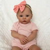 A newborn reborn baby doll with light skin, blue eyes, and pink clothing, wearing a peach bow.