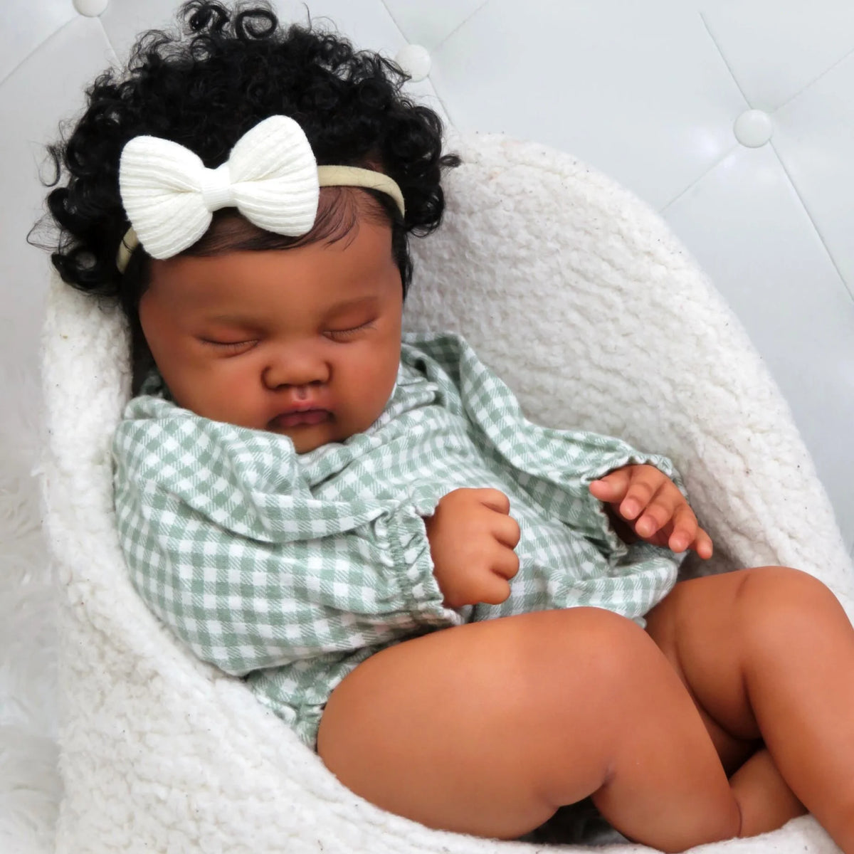 A newborn baby with curly hair wearing a green gingham outfit and a white bow, resting in a white knitted basket.