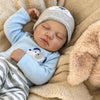 A newborn vinyl reborn baby doll wearing a gray knitted hat and light blue long sleeve shirt, resting on a soft beige blanket