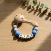 A wooden pacifier clip featuring a silicone charm spelling out BYBA