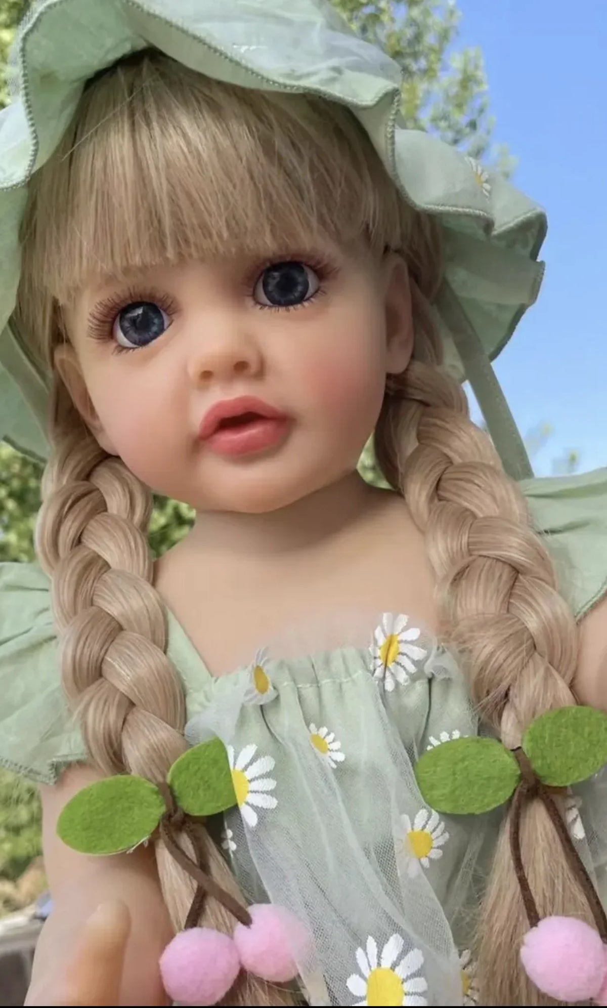 A close-up of a blonde toddler doll with long braided hair wearing a light green dress and a matching bonnet.