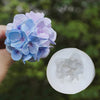A hand holds a purple blue silicone flower blossom mold with delicate petals against a blurred green background.