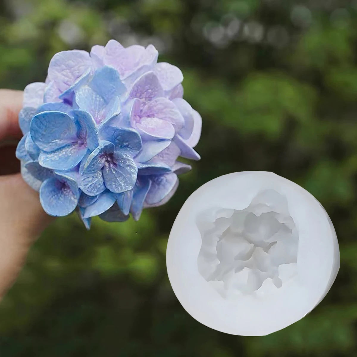 A hand holds a purple blue silicone flower blossom mold with delicate petals against a blurred green background.