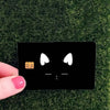 A black card with a cat face design featuring white eyes and a thumbs-up hand gesture