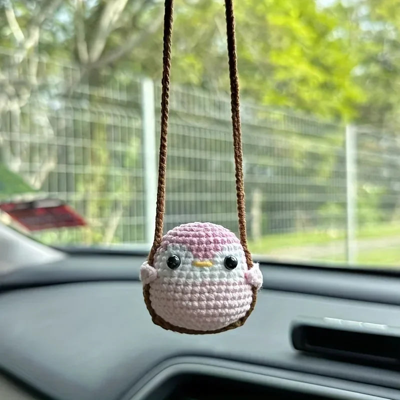 A handmade crochet chicken charm with pink and white features is hanging from a string inside a car.