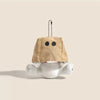 A tan plush charm shaped like a head with two black dots, attached to a chain, resting on a white fluffy object.