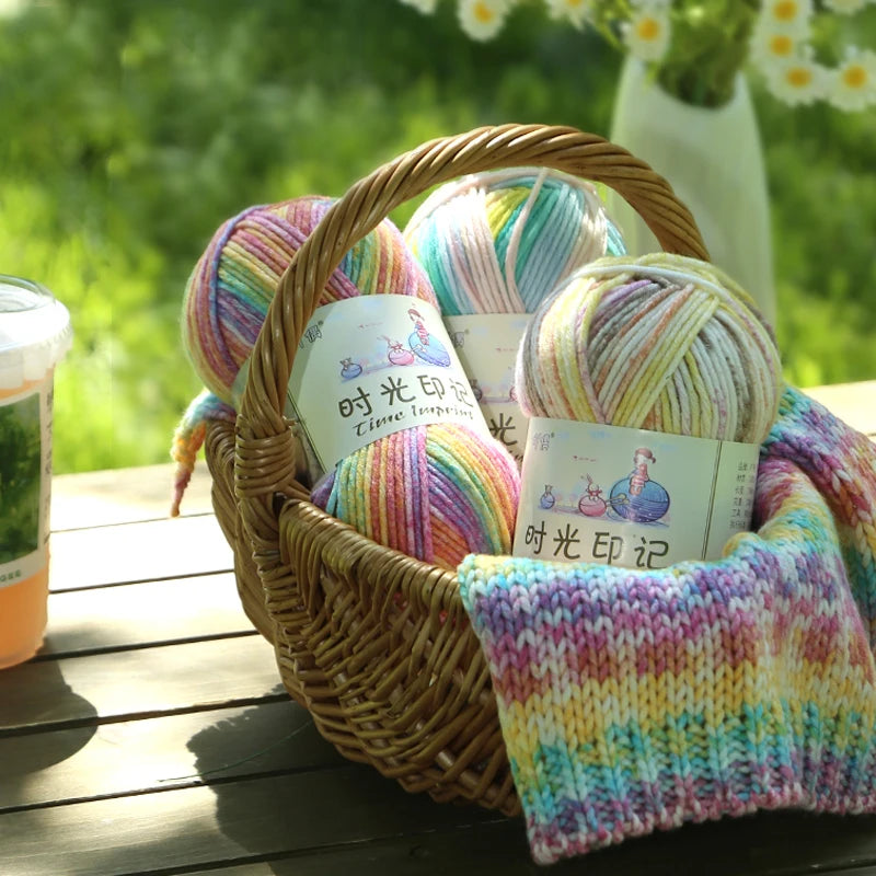A woven basket filled with three balls of gradient acrylic yarn in various colors, resting on a wooden surface.