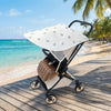 A beige and white lightweight UV resistant sunshade for a baby stroller is placed on a wooden deck near the ocean.
