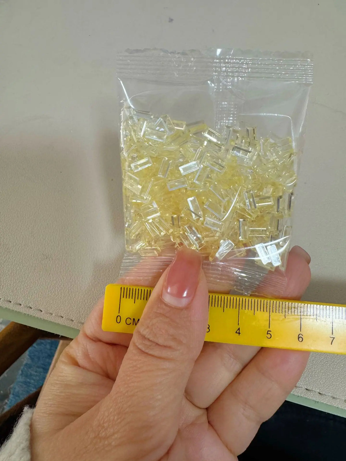 A hand holds a clear plastic bag filled with yellow acrylic ice strips for nail art and DIY crafts next to a ruler.
