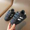 A hand holds a black Adidas sneaker with white stripes and hook-and-loop closure on a beige surface.