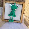 Handmade crochet frog hanging ornament with green body and large eyes displayed in a wooden frame