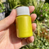 A hand holds a small bottle of metallic gold acrylic paint for crafts against a blurred green outdoor background.