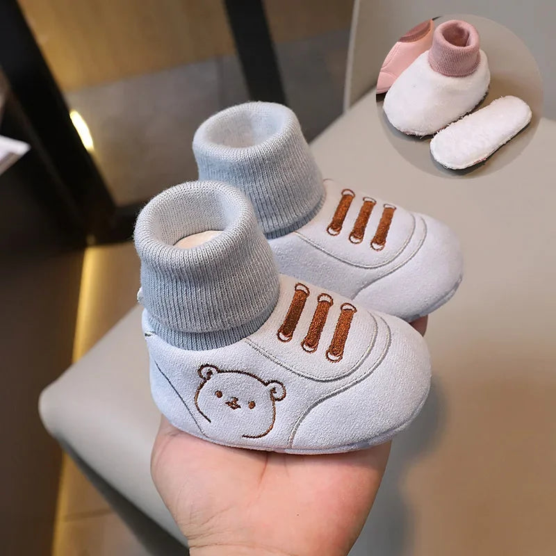 A pair of light gray knit bear booties for a baby, shown resting on a hand with other similar items.