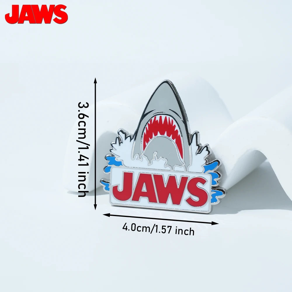 Silver enamel lapel pin featuring a shark tooth design and the word JAWS on a white background.