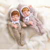 A baby doll wearing a cozy gray knitted romper outfit with a white hood and bow, lying on a fluffy white surface.