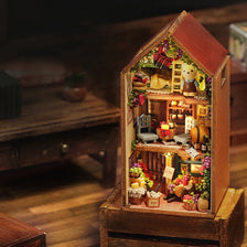 A wooden miniature dollhouse kit featuring a detailed diorama of a cozy, rustic interior with small figures and decorative el