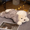 A soft, lying dog plush toy with gray and cream fur resting on a gray fabric surface.