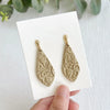 A pair of gold-toned earrings with a leaf shape and floral texture on a white card.