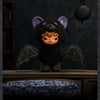 A dark plush doll with purple ears and orange hair has black wings, displayed on a wooden surface.