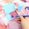 A hand uses a blue pen to apply dots of paint onto a diamond painting canvas inside an open blue box.
