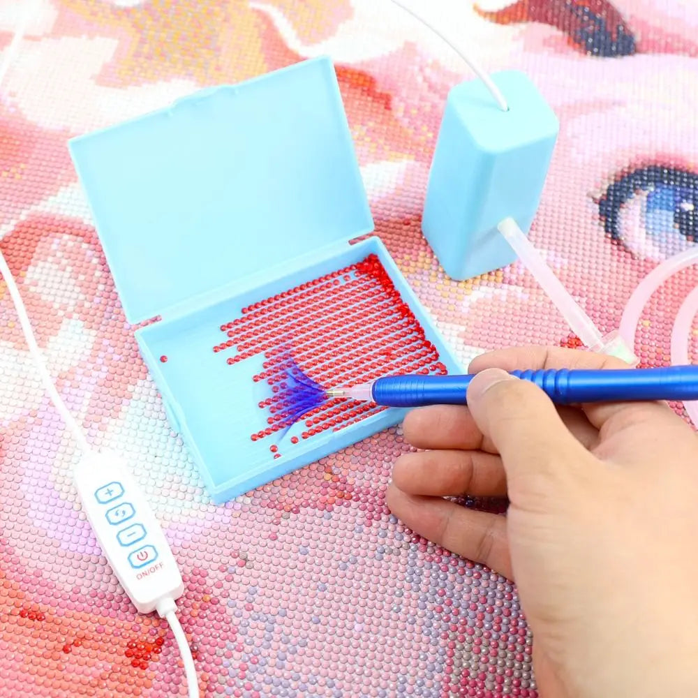 A hand uses a blue pen to apply dots of paint onto a diamond painting canvas inside an open blue box.