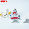 A silver enamel lapel pin featuring the word JAWS with a stylized shark tooth design.