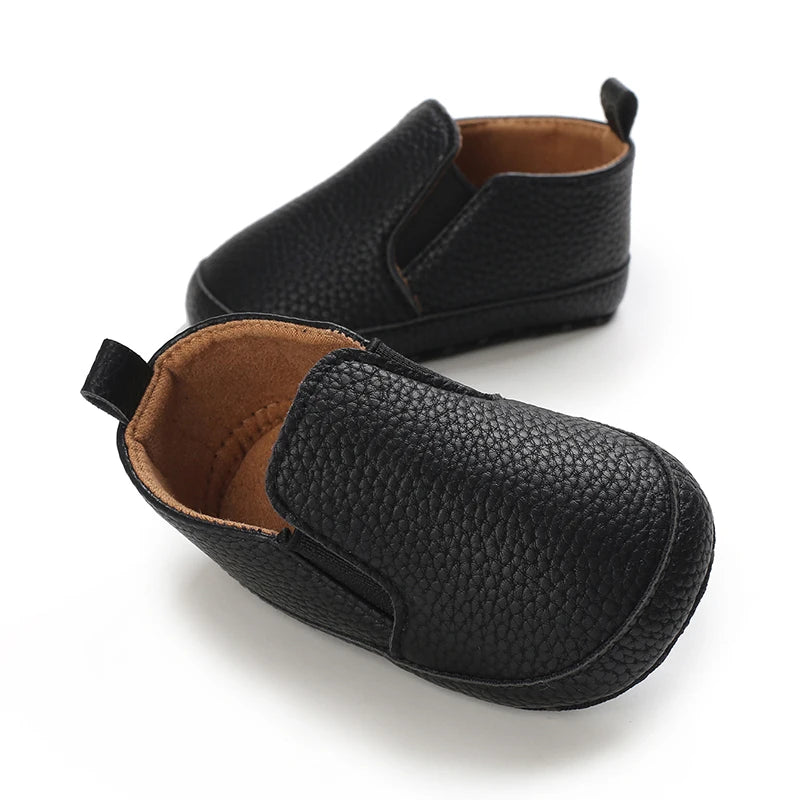 A pair of black baby first walker sneakers with a dinosaur print detail shown from the side.