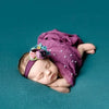 A newborn baby wearing a purple knit wrap and a floral pearl headband, lying on a teal background.