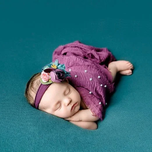 A newborn baby wearing a purple knit wrap and a floral pearl headband, lying on a teal background.