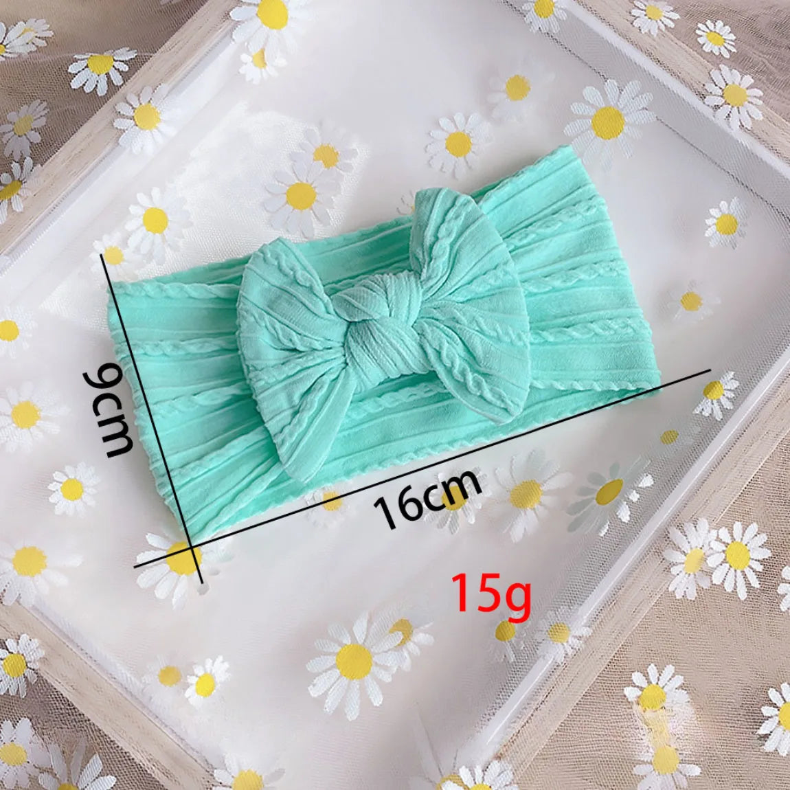 A light mint green fabric bow baby headband is displayed on a white surface with scattered daisy flowers.
