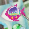 Leuchtend pinkes Fisch-Sticker mit klarem Kuppelinneren und gelben Akzenten wird mit Pinzette gehalten.