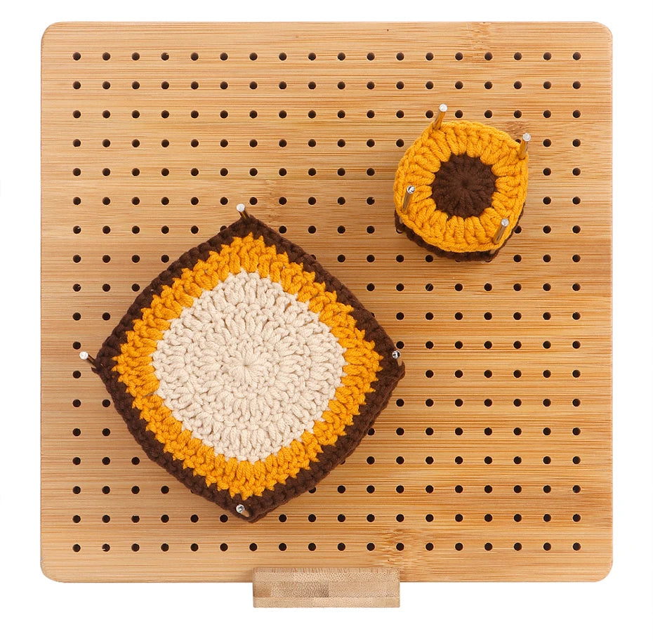 A yellow and brown crochet object is placed on a bamboo blocking board with pins and a wooden stand.