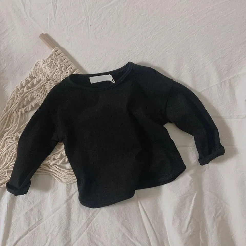 A black long sleeve cotton tee is laid on a white surface next to a cream-colored knitted item.