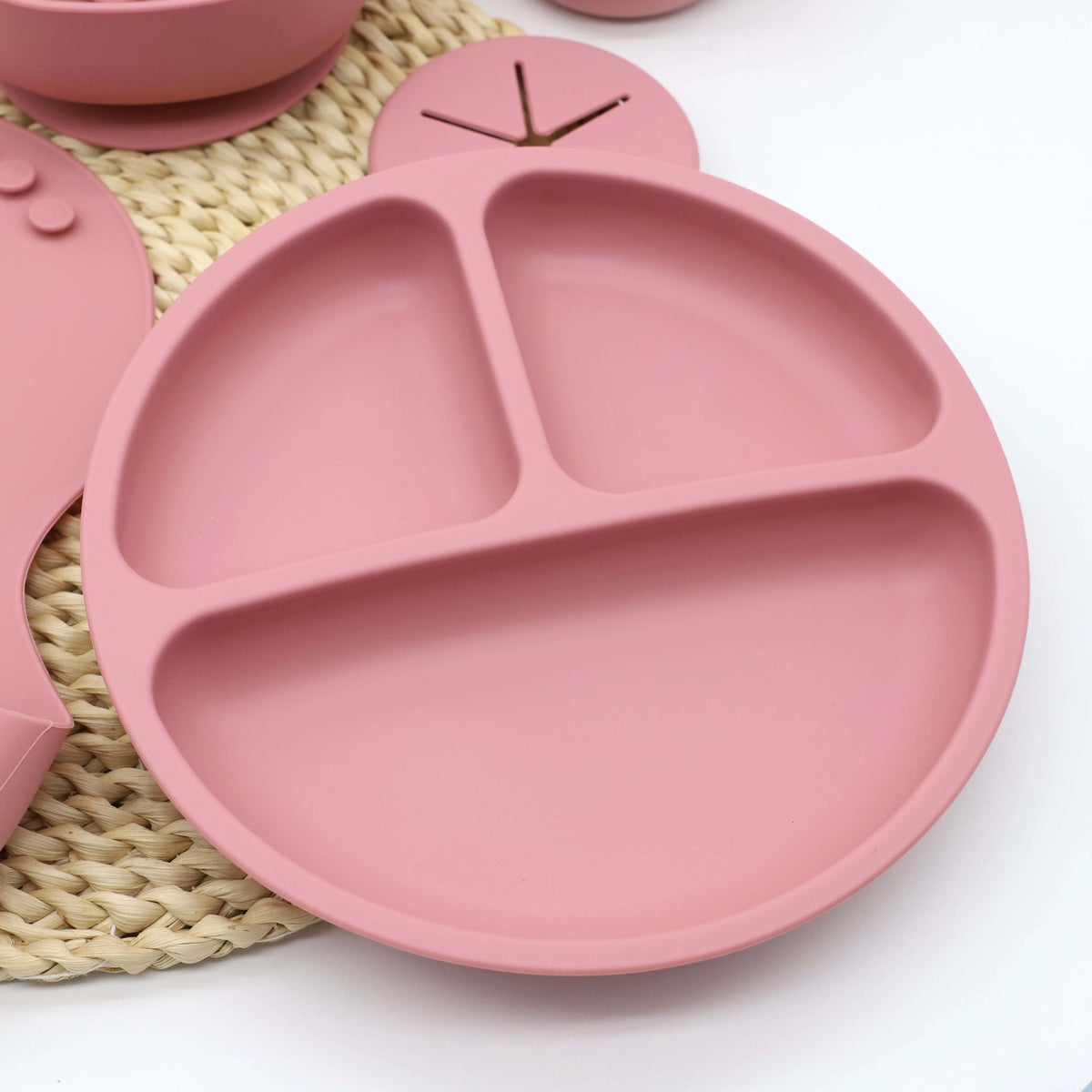 A pink silicone baby tableware set with suction cups is shown resting on a woven mat.