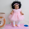 A pink reborn toddler doll with hand-rooted black hair wearing a ruffled dress and white tights.