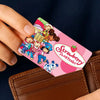 A hand holds a pink and green cartoon credit card sticker set featuring Strawberry Shortcake characters on a brown leather wa