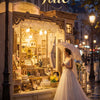 A woman in a white gown stands before a detailed display of jewelry and ornaments, set within a vintage-style shop window.