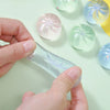Hands hold a clear plastic steamer basket containing several translucent squishy toys.