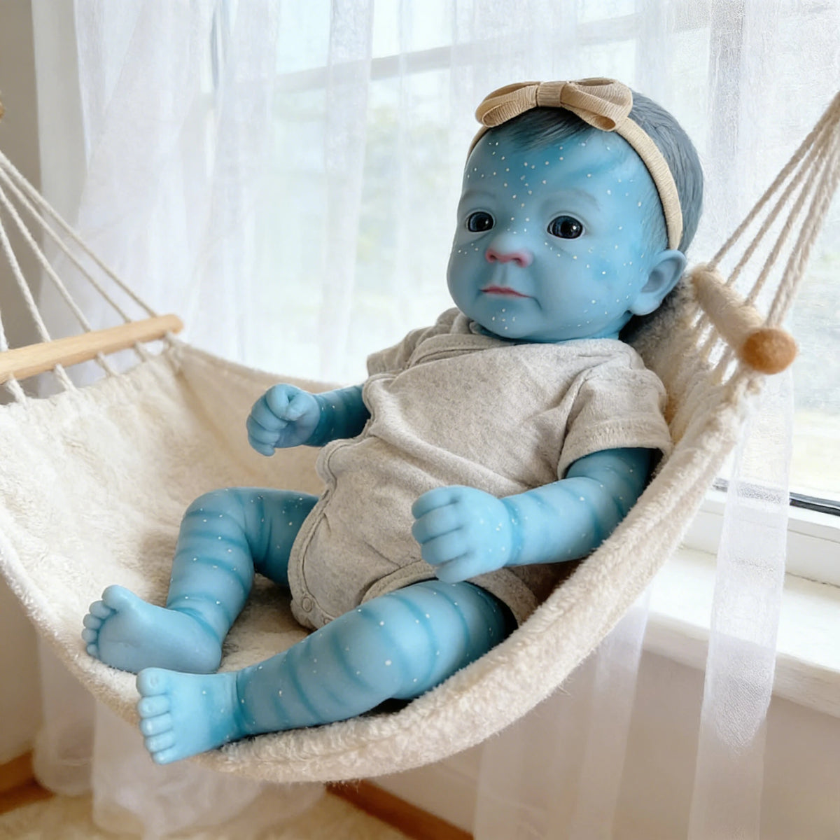 A blue reborn baby doll with a headband is seated in a white hammock.
