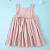 A pink linen jumper dress for a toddler girl with a gathered waist and long tie details, set against a light blue background.