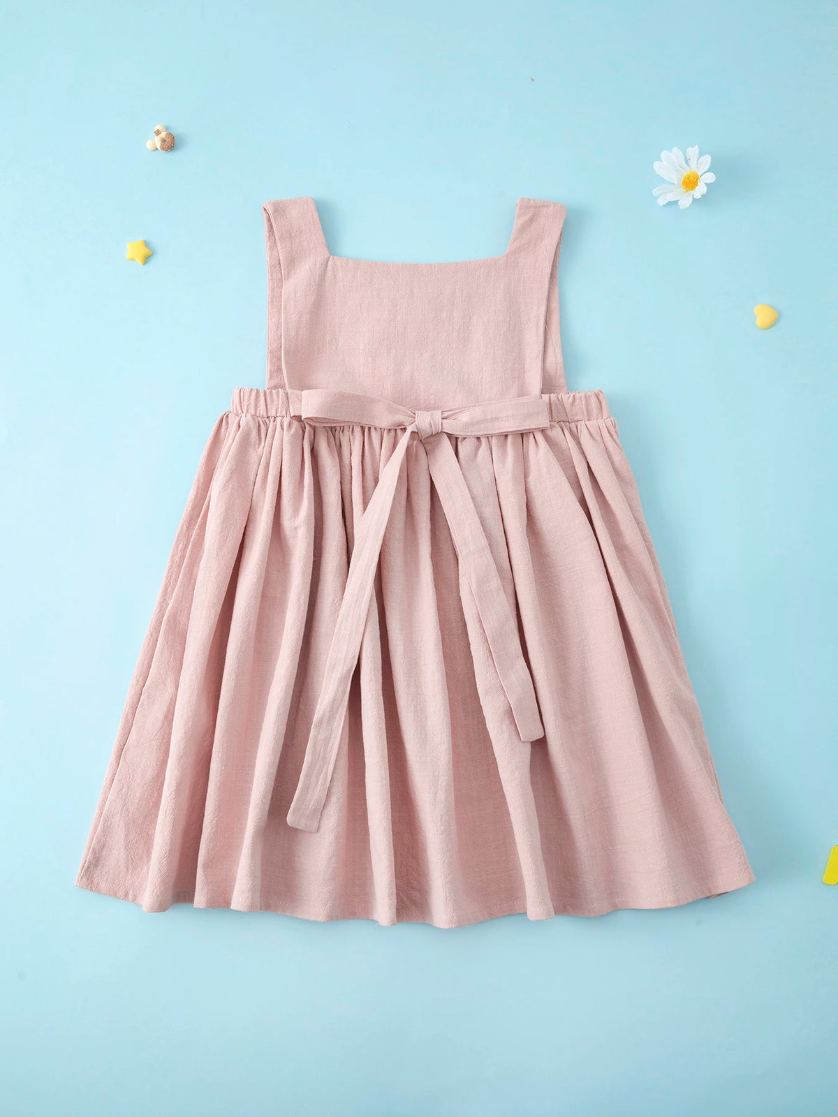A pink linen jumper dress for a toddler girl with a gathered waist and long tie details, set against a light blue background.