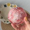 A hand holds a translucent pink stress ball with glitter and floral inclusions against a light background.