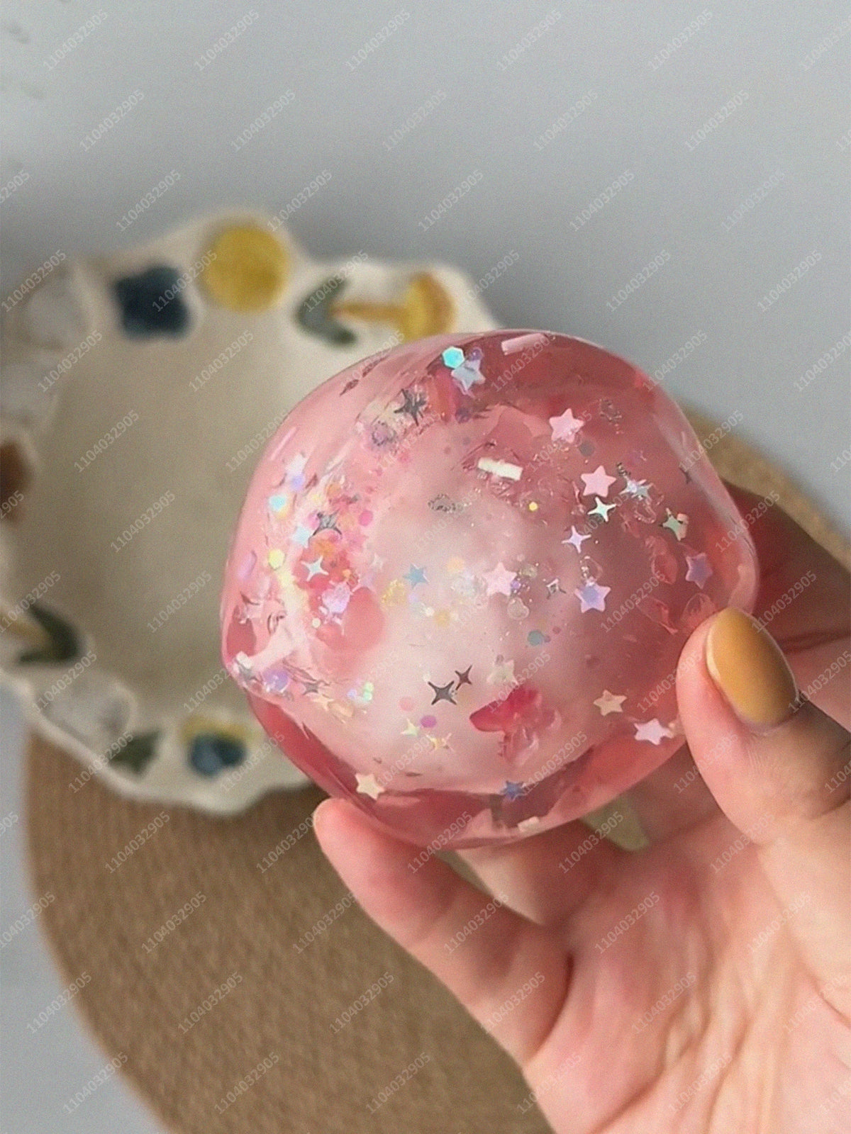 A hand holds a translucent pink stress ball with glitter and floral inclusions against a light background.