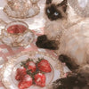 A fluffy black and white cat sits next to a tea set and a plate of strawberries on a pink checkered surface.
