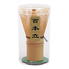 A clear glass container holding a traditional bamboo matcha whisk set with a wooden base.